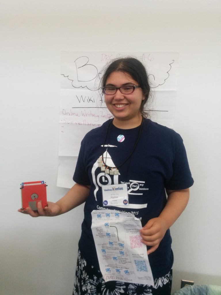 Alyssa Hillary stands before a DRC Wiki Topic brainstorming poster with a list of possible topics and DRC Wiki Quest SWAG, including the floppy disk pencil holder and with the DRC Wiki Quest game sheet