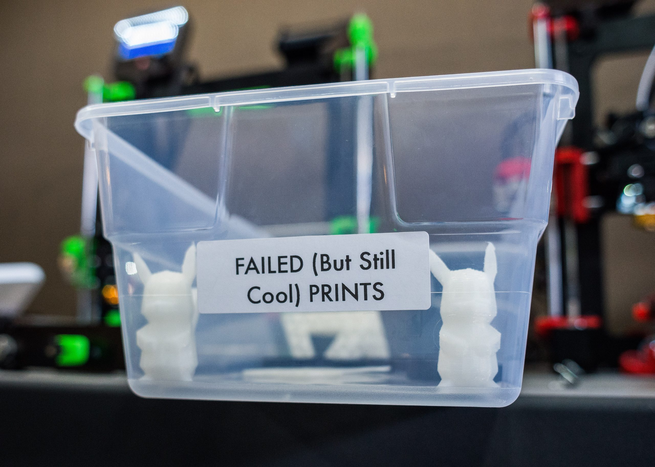 A clear plastic box with the label "Failed (but still cool) prints" contains small 3D printed objects and sits in front of two 3D printers.