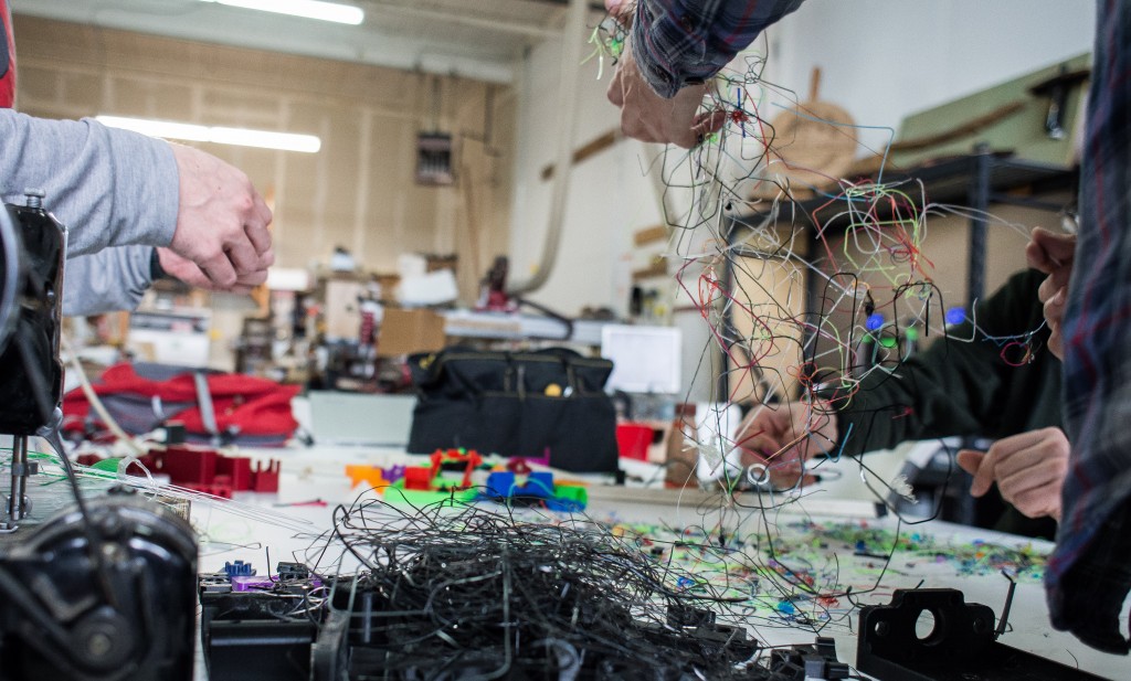 Three sets of hands are pictured sorting through a mass of tangled, multicolored, strands of 3D printer filament.