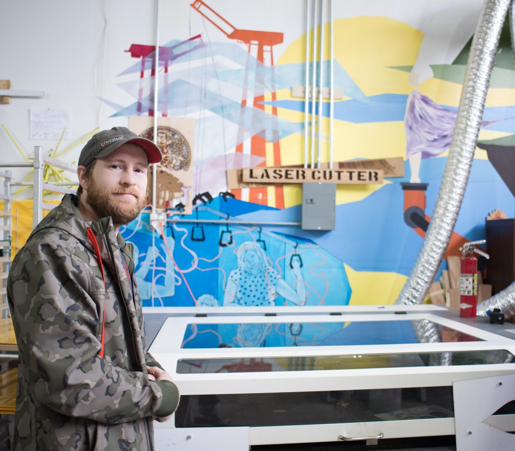 Eric Renn, founder of SoDo MakerSpace, wears a jacket and cap, and he stands in front of a large laser cutter, in front of a wall painted with a multi-colored mural.