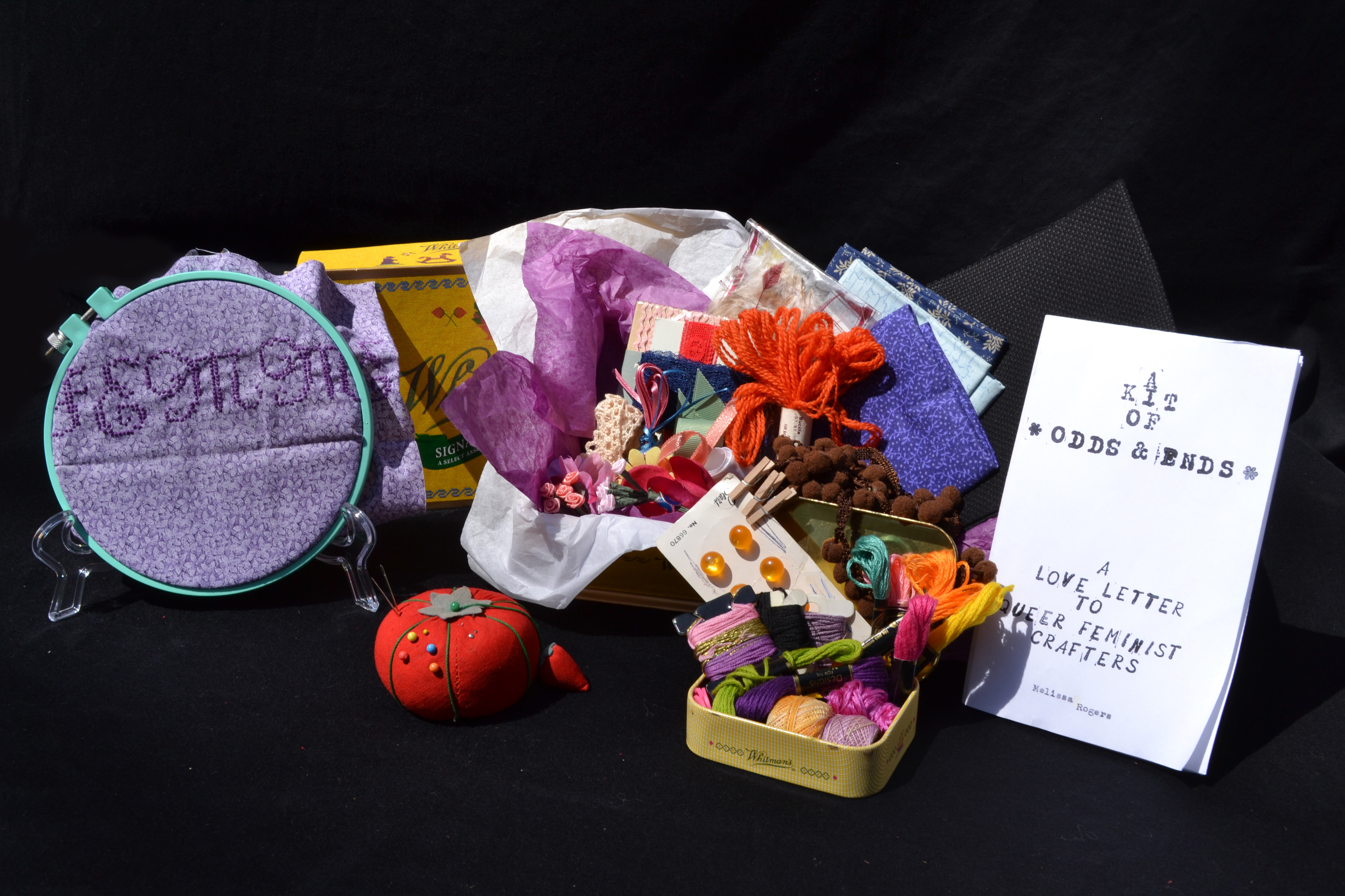 Contents of the kit arranged against a black backdrop including an embroidery hoop, a pincushion, the zine, and a large and small box stuffed with craft materials