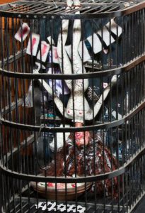 A Barbie doll hangs upside down within a birdcage. The Barbie is mangled and appears her body has taken a toll from the numerous medical procedures that she has been forced to endure.