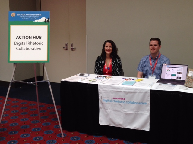 Two people sit behind a table covered by pamphlets and flyers. On the left is Sara West and on the right is David Coad. To the left of the table, a sign for the Action Hub reads "Digital Rhetoric Collaborative"