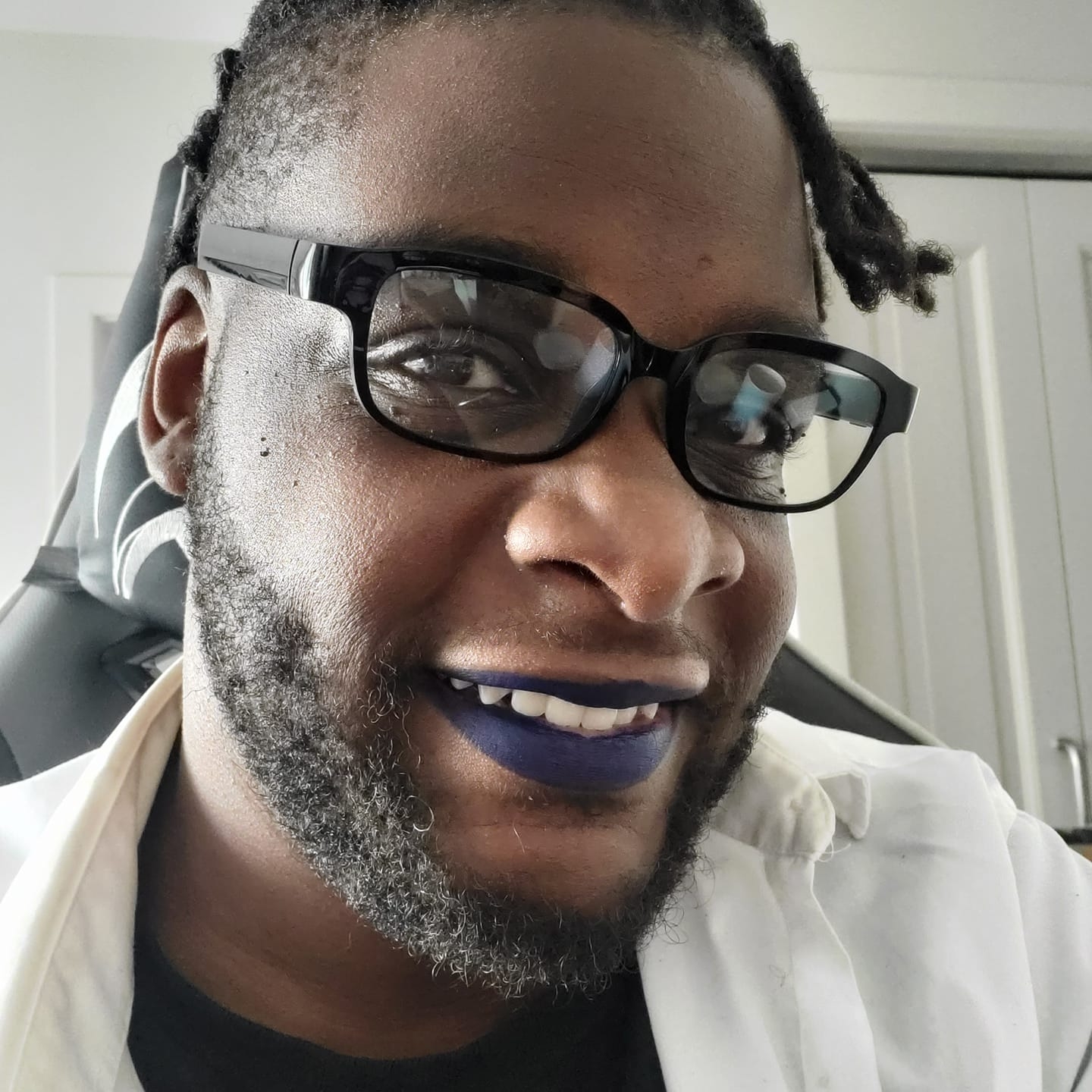 A picture of a black man with a shaved head and dreadlocks wearing glasses, and dark blue lipstick