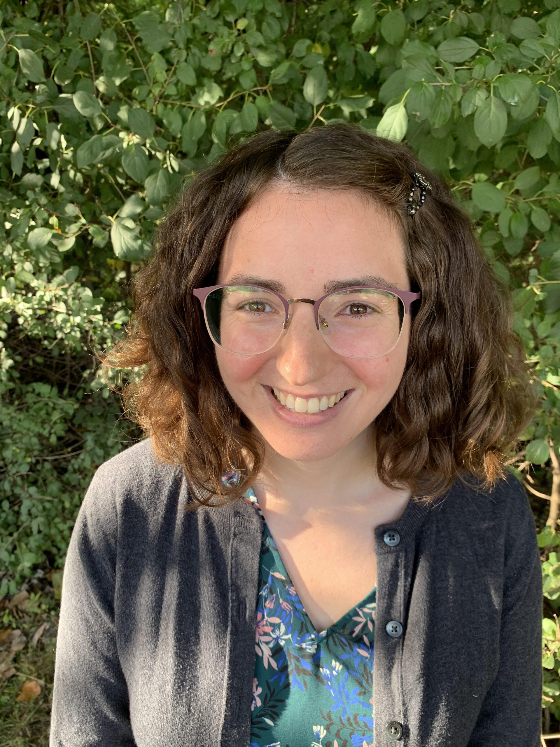 A woman with chin-length hair and glasses standing in front of greenery