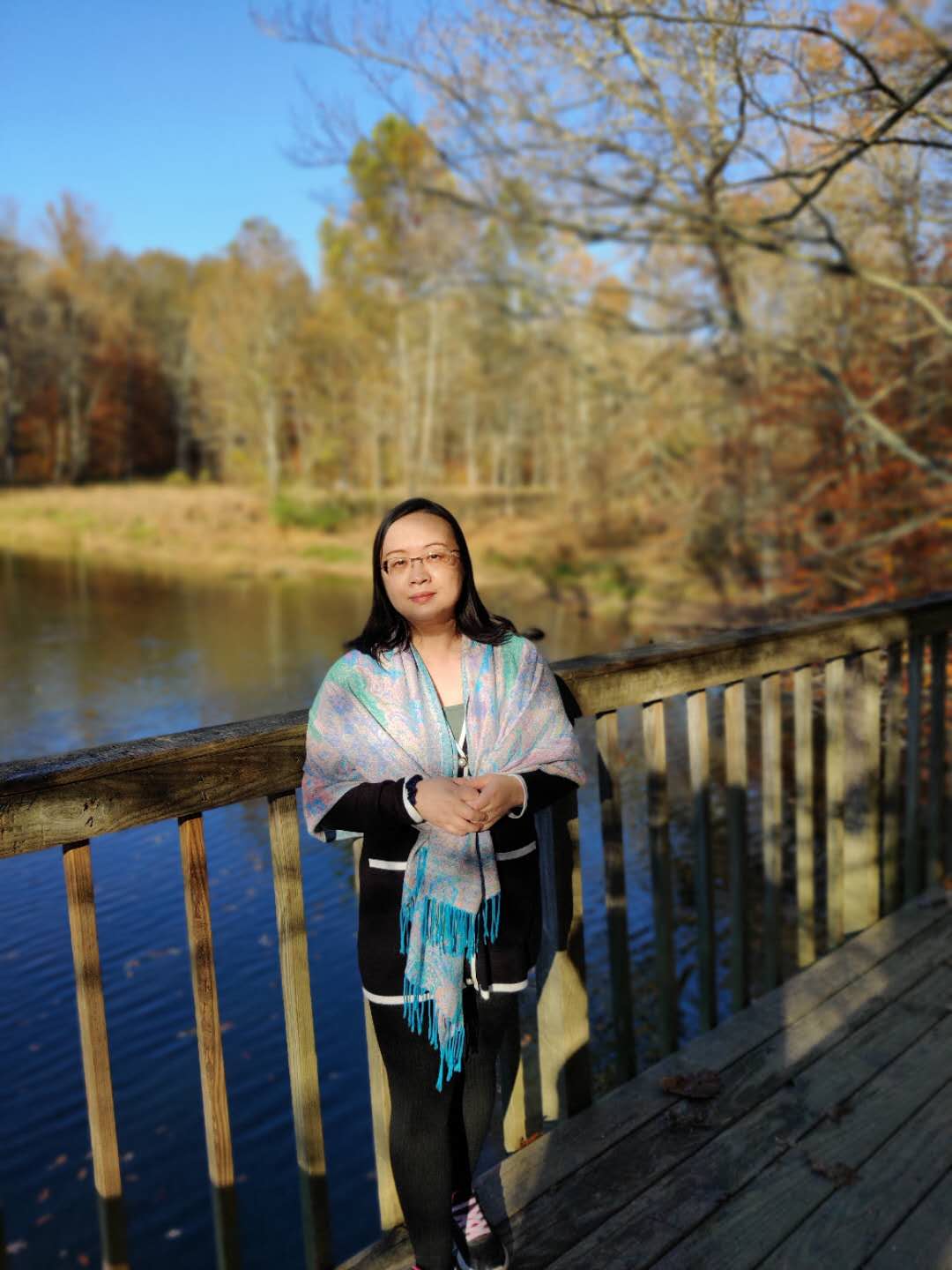 A woman leaning against the rail of a bridge with woods, a lake, and blue sky as the background