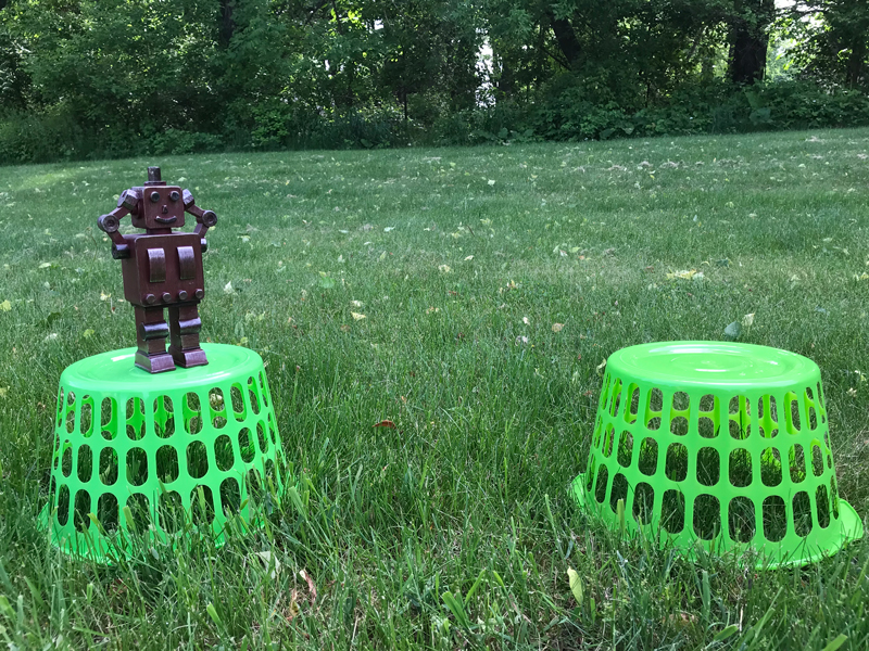 A confused robot toy is pictured on top of an upside-down green basket. Another green basket is about a foot away. Robot's hands are on its head since it cannot cross the gap.