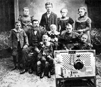 Black and white image of men, women, and children in old, possibly nineteenth century, clothes watching a TV.