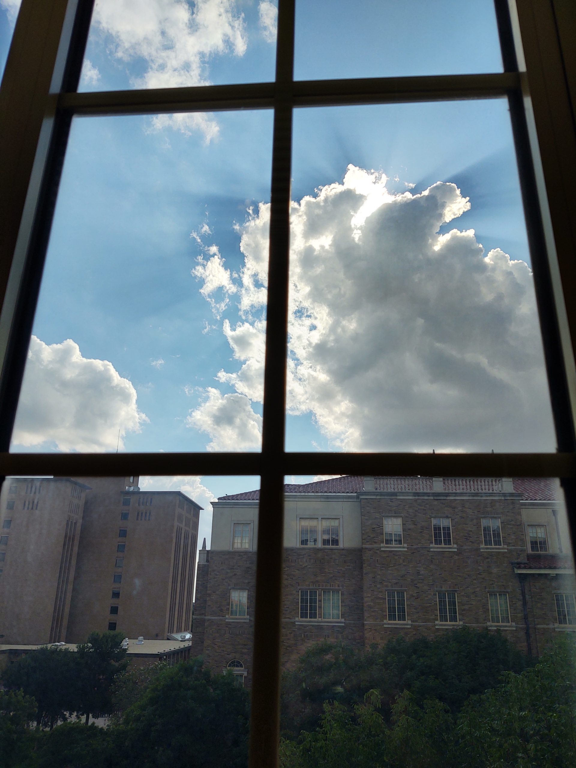 Picture is of sun shining from behind clouds with campus buildings and trees also visible below. 